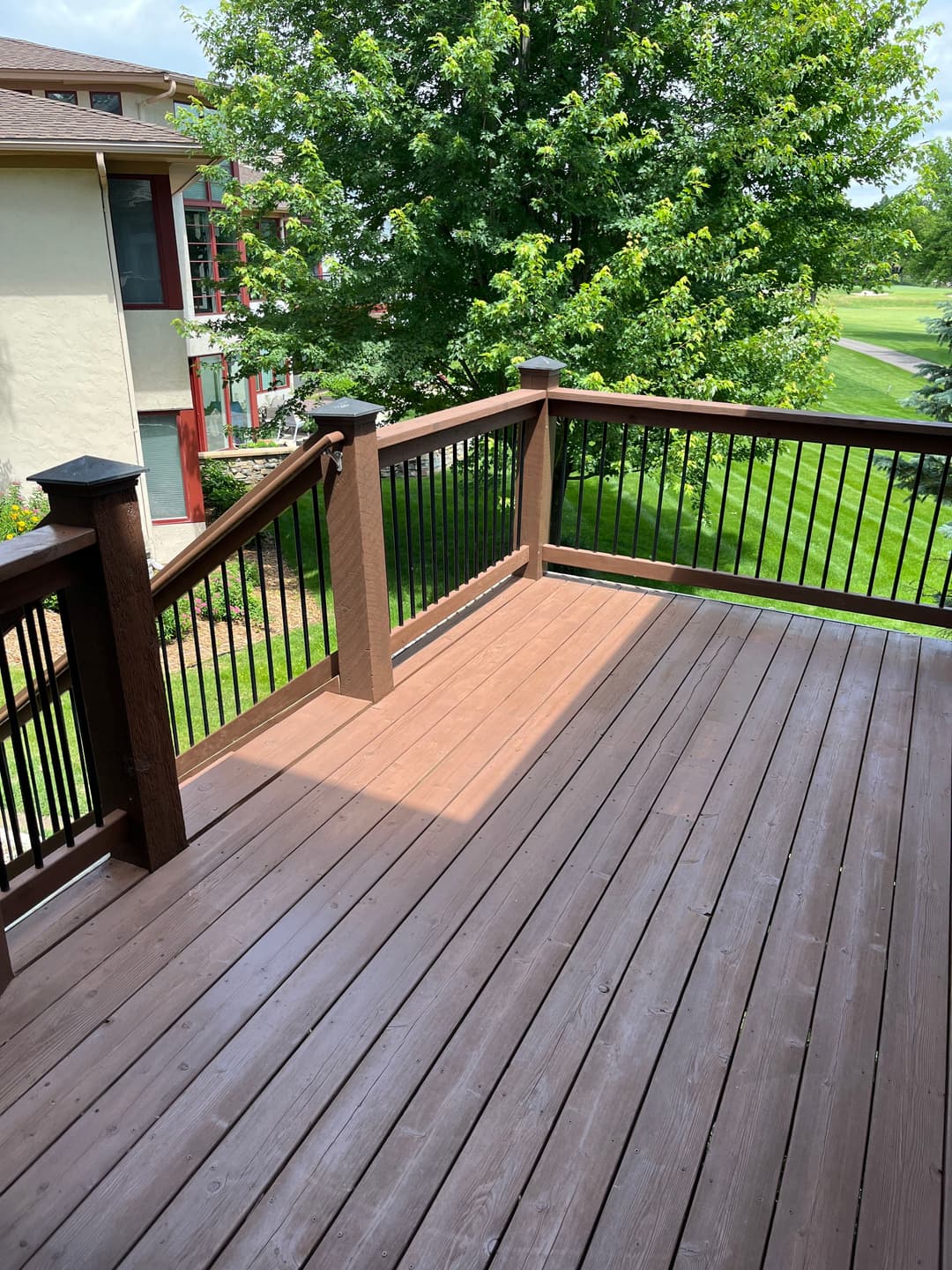 Spacious brown deck with railing overlooking green lawn and trees on a sunny day.