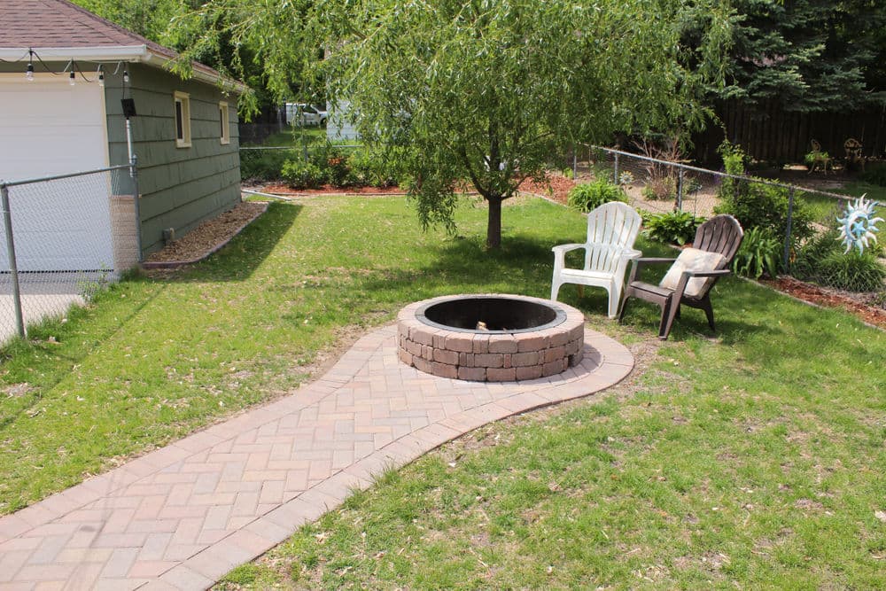 backyard fire pit with brick circle, paved walkway, and two chairs in a green lawn setting