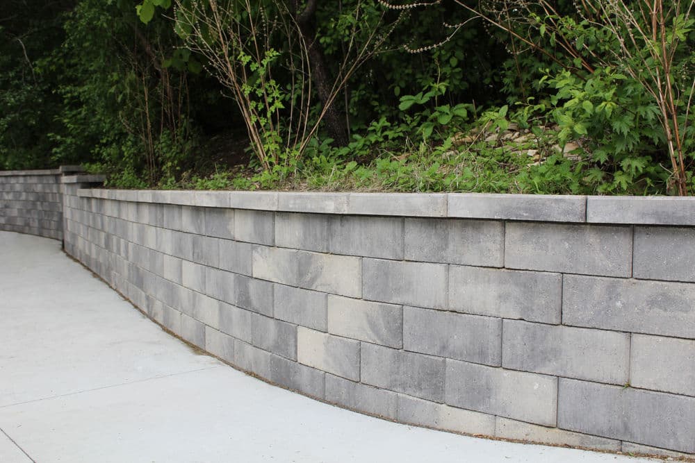 Curved gray stone retaining wall with greenery against a path in a landscaped area.